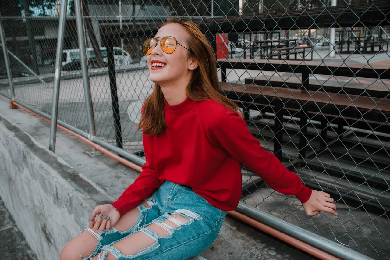 A fashionable woman in sunglasses and tattered jeans smiles outdoors by a chain link fence.